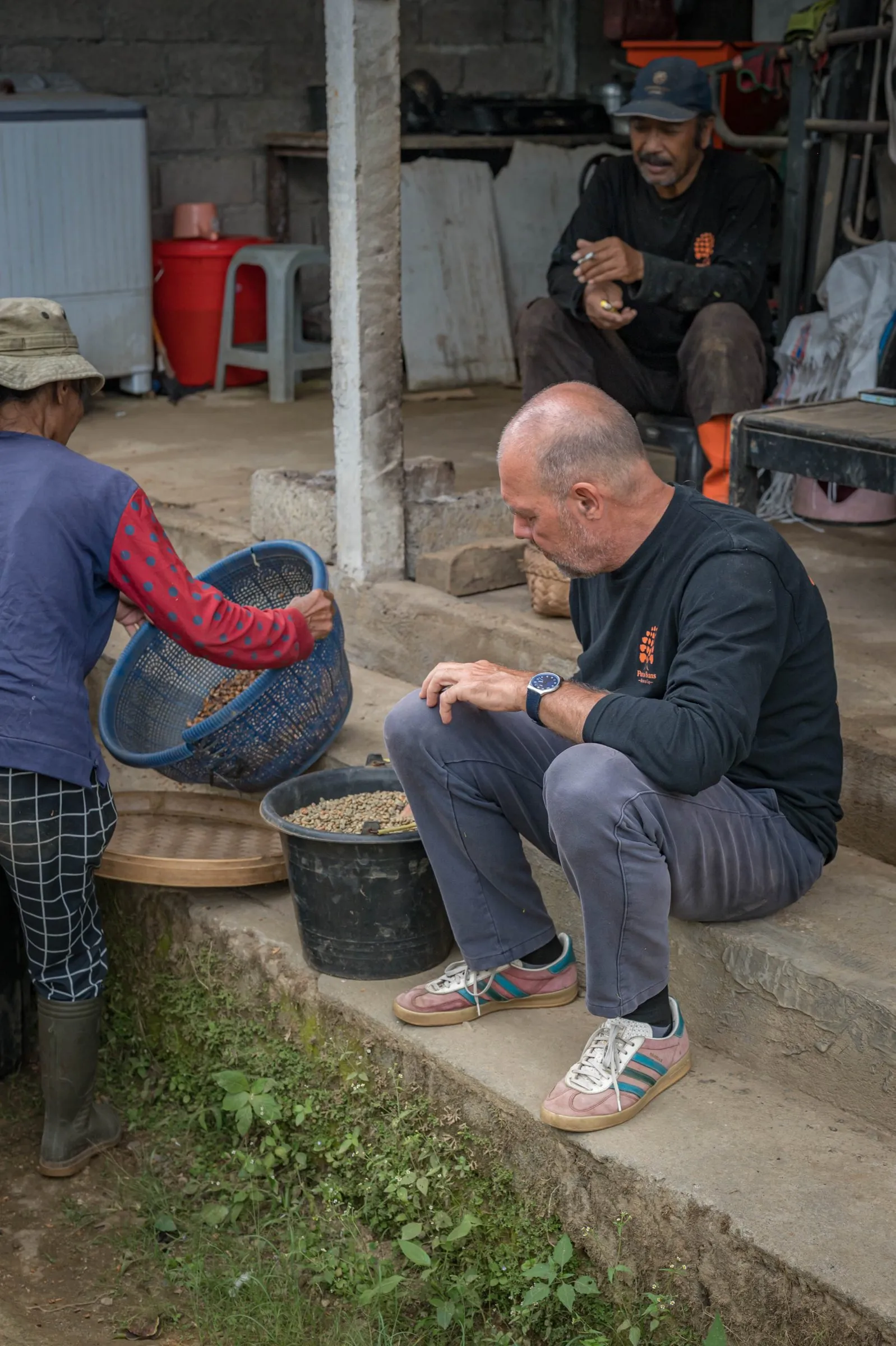 Ibu Ketut pouring parchment coffee from a basket, with a man in a Portus Beans shirt on the step